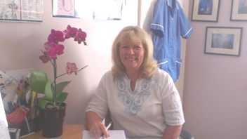 Beverley Moss sitting at a small table in her treatment room, in a white top and blue patterned scarf, with a pink orchid on the table beside her and a pale blue towel on the wall behind.